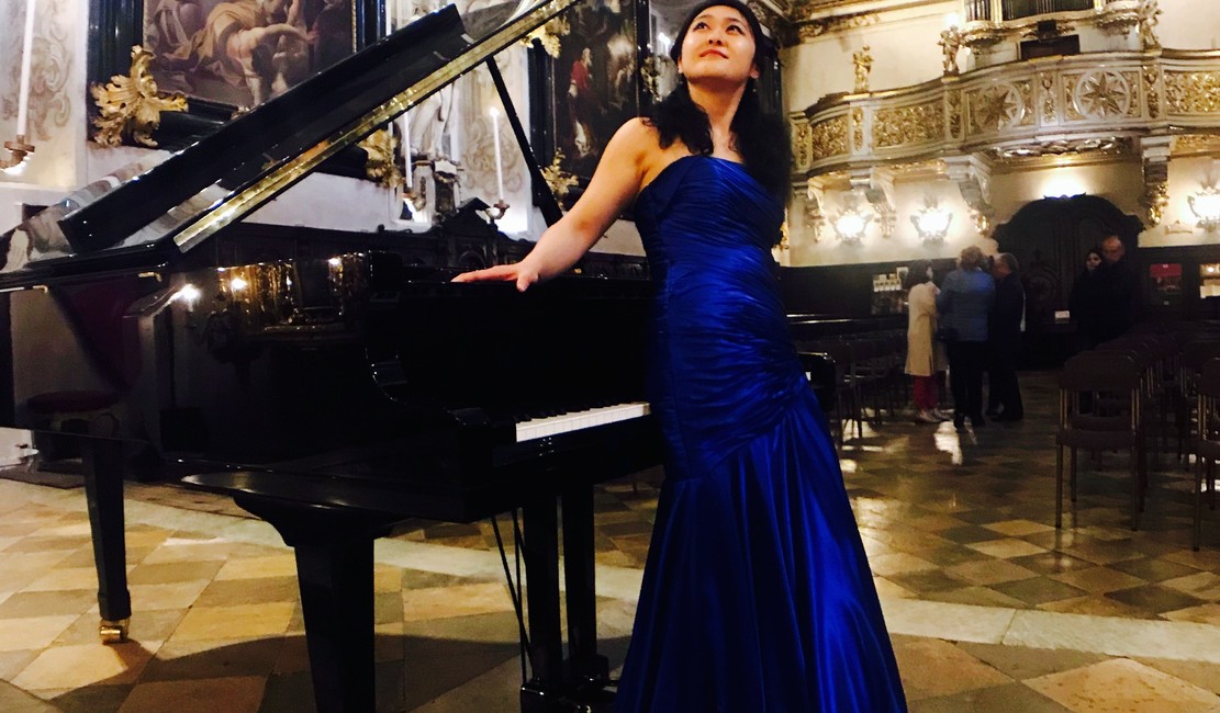 Lishan Xue standing by a grand piano in a blue dress.